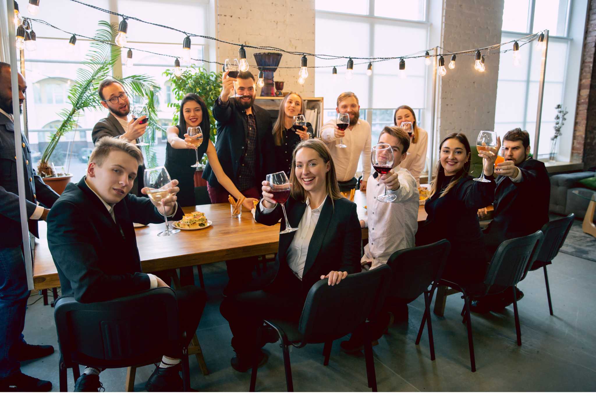 Group of professionally dressed colleagues raising wine glasses in a celebratory toast around a modern office dining table, with string lights and snacks creating a festive atmosphere.