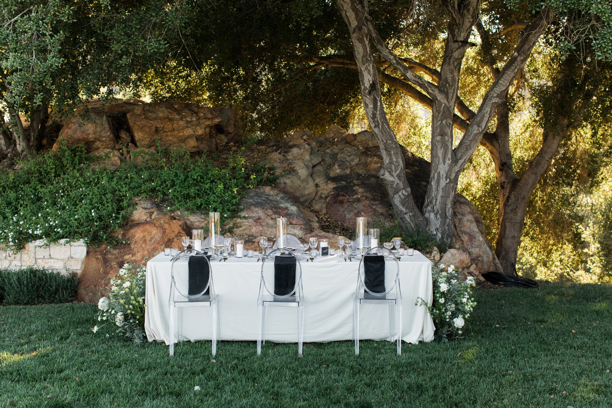 Outdoor Event Tablescape with White Linen and Candles Outdoor table beneath trees with white linen, clear chairs, black accents, candles, and white flowers.